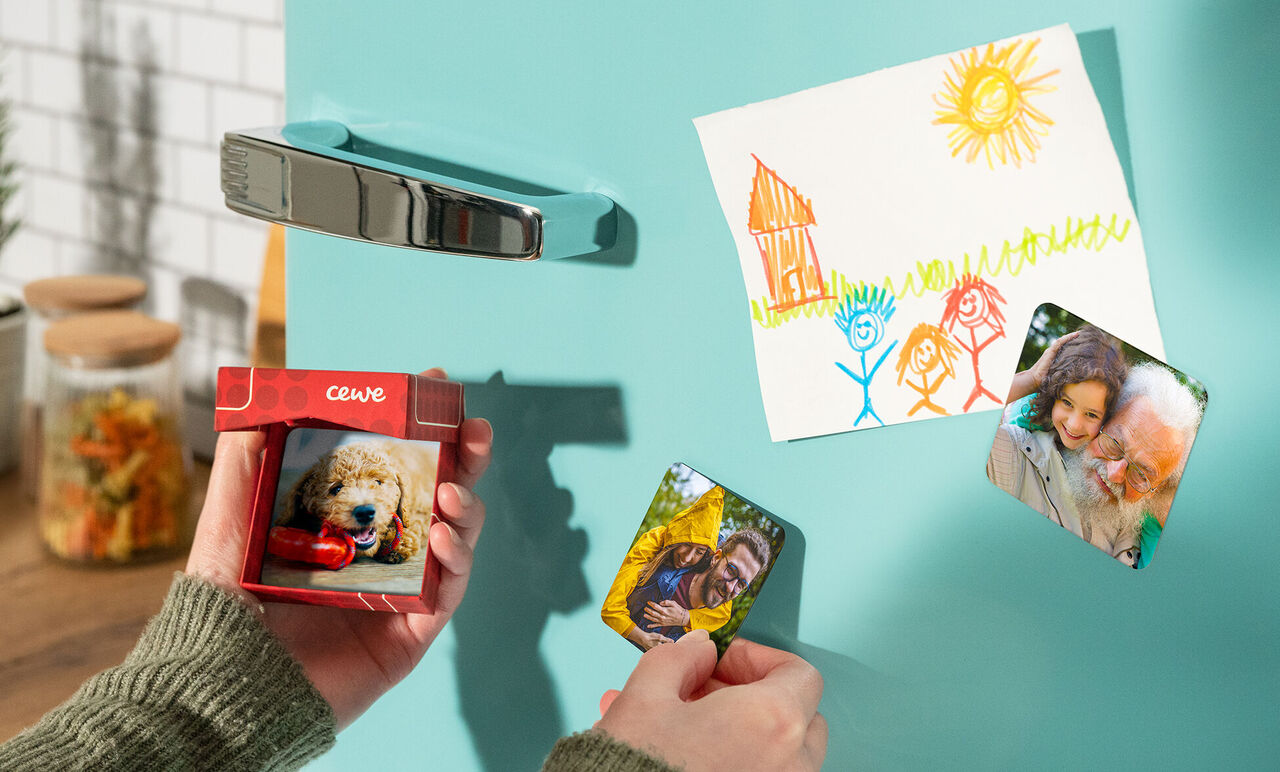 Hand holds a small CEWE greeting card with a dog picture, a drawing hangs on the fridge door.