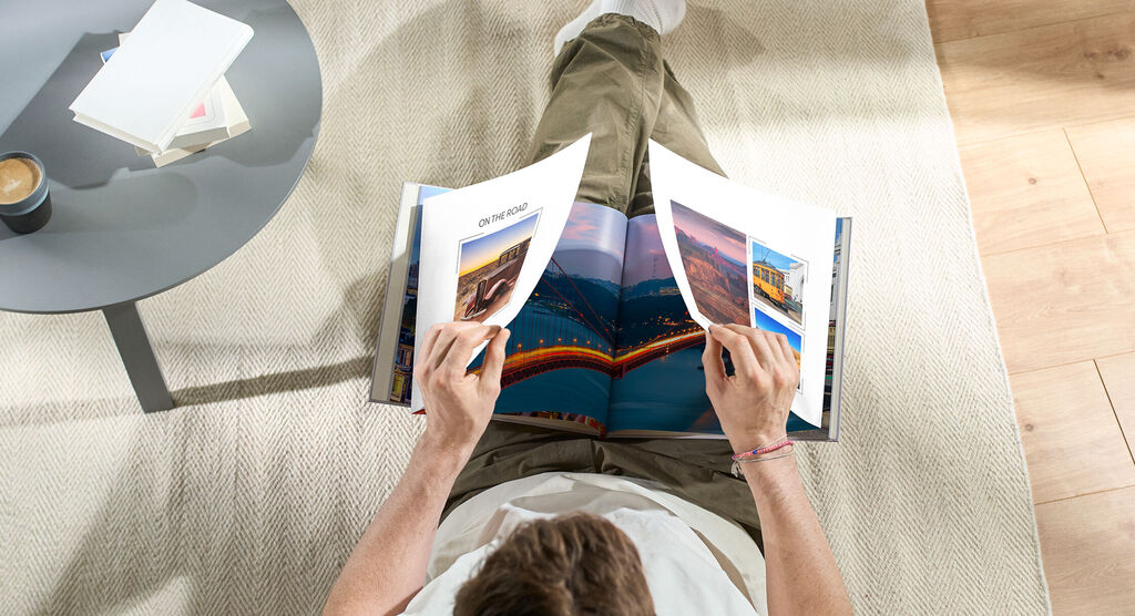 A person flipping through an open photo book on the floor, surrounded by a table with a cup.