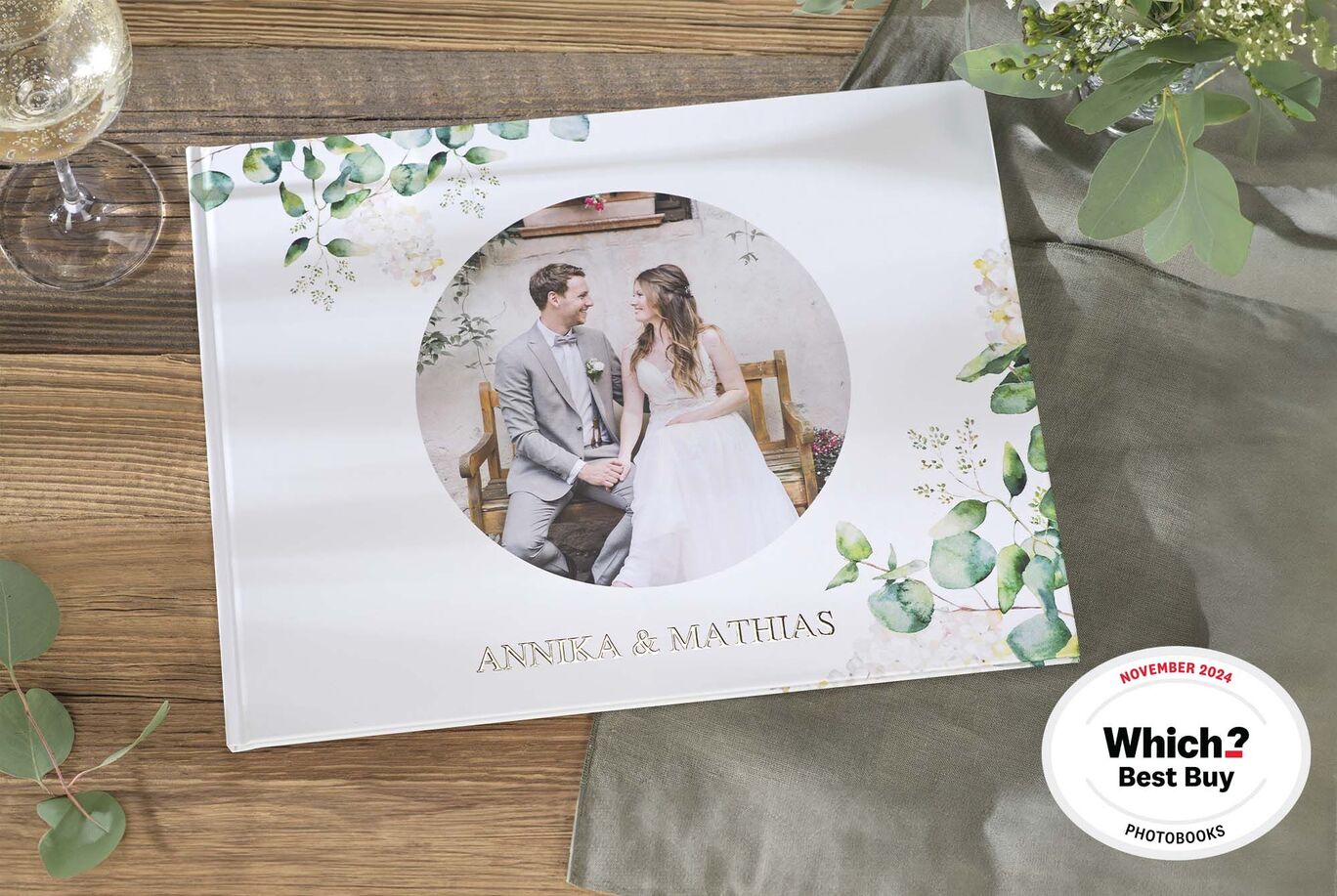 A CEWE photo book with a wedding photo of Annika and Matthias. The album has a floral frame and lies on a table next to a glass of sparkling wine. The sticker shows it was awarded.