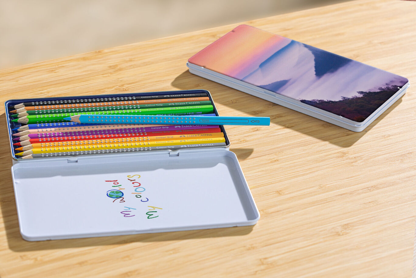 Colored pencils in a writing box with personal design next to a notebook on a wooden table.