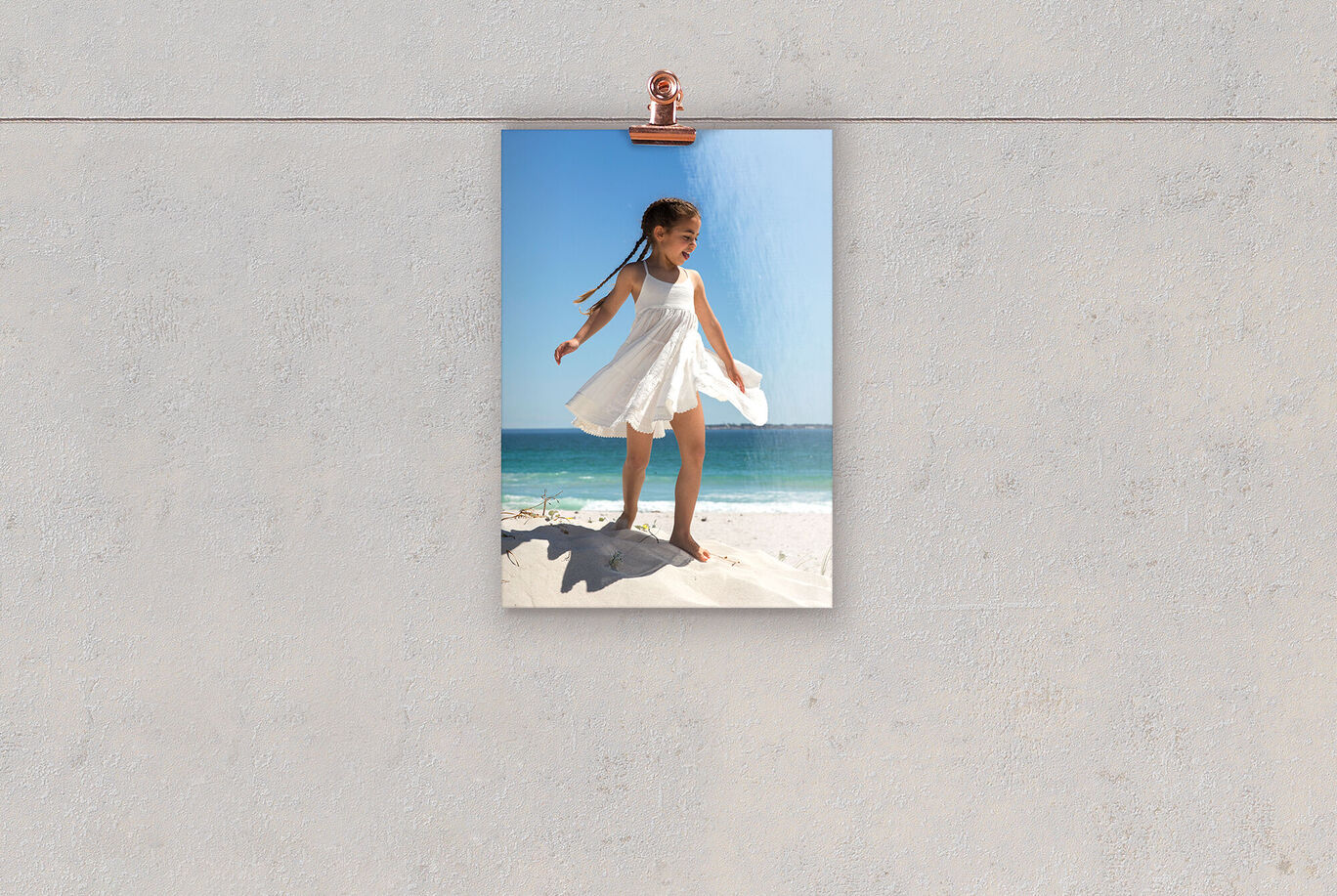 A happy girl in a white dress stands on a sand dune at the beach. Behind her are the blue sea and sky.