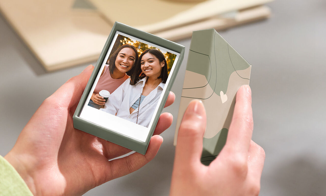 A person holds a green gift box with a photo print in hand, surrounded by soft colors