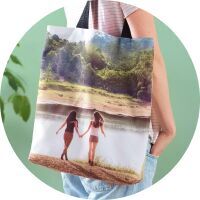 Two friends hold hands at the beach, carrying a personalized CEWE photo bag with their shared picture.
