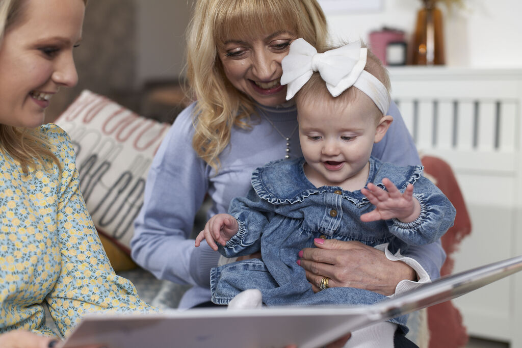 Child with a picture book on mother's lap. They sit cozily in a living room.