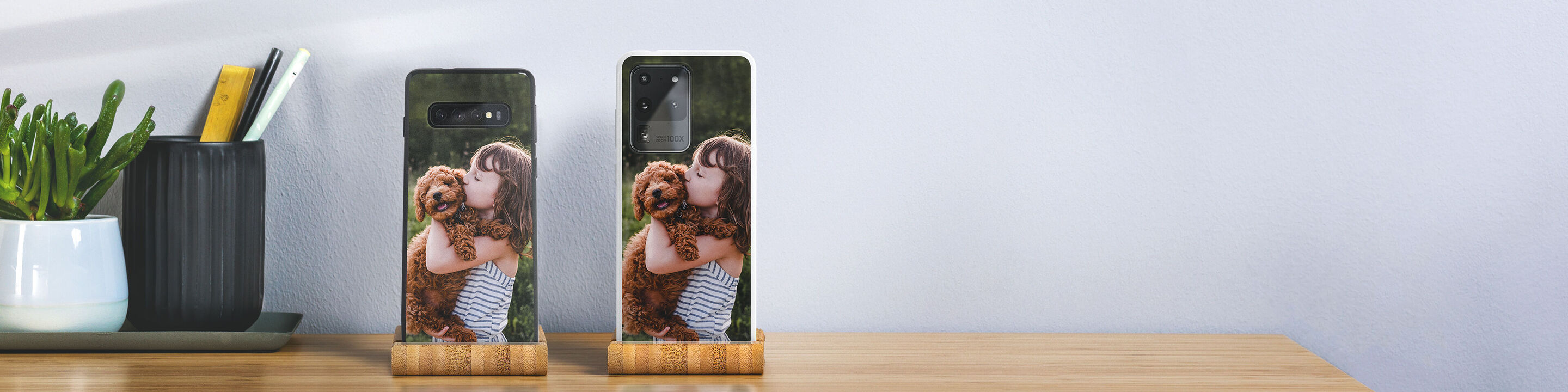 Two personalized smartphone cases with images of a girl and her dog. They are on a table next to a plant.