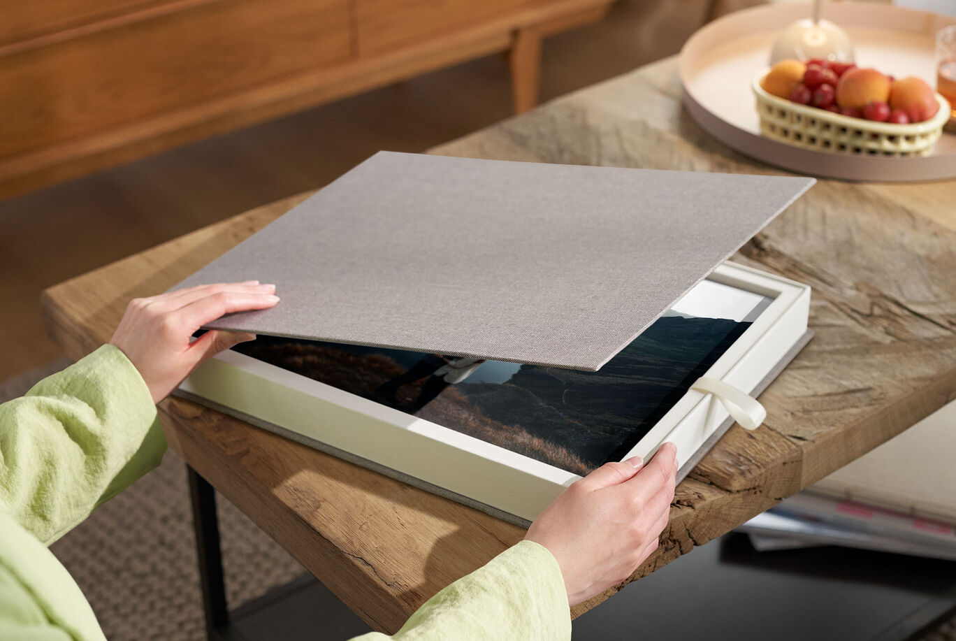 A person lifts the cover of a CEWE photo book on a wooden-patterned table.