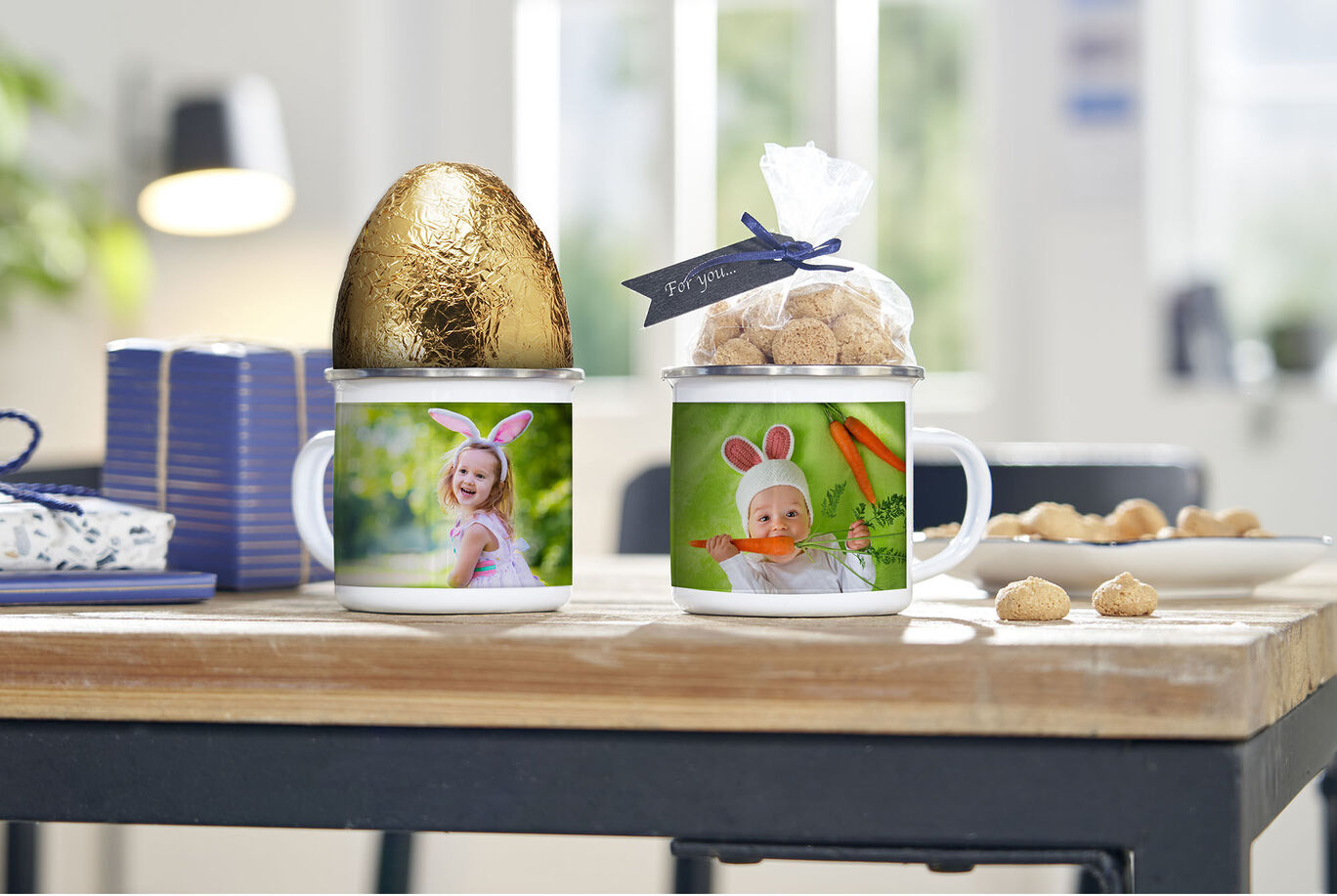 Two personalized mugs on a table, decorated with a golden egg and candies in bags.