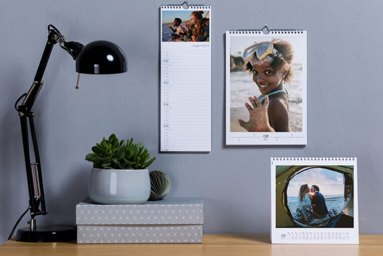 Two wall calendars with personal photos, one shows a beach scene and the other a couple on holiday. A green plant is on the table along with a desk lamp.