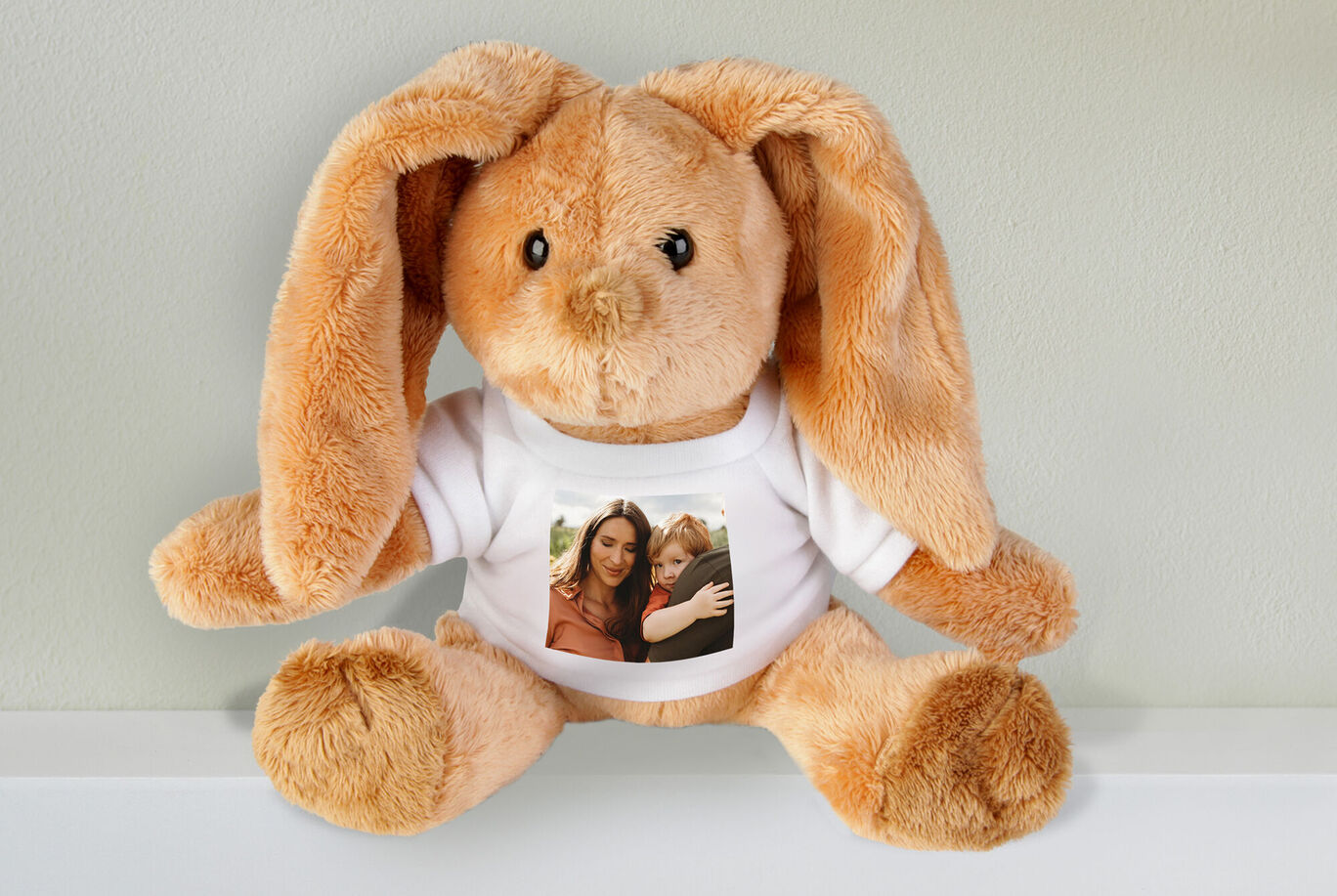 A cute stuffed rabbit wearing a T-shirt with a photo. The rabbit sits on a shelf and radiates joy.