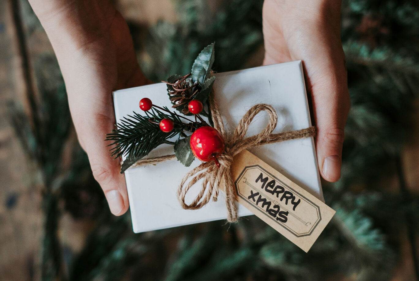 A hand holds a lovingly wrapped gift with a cord and a Christmas tag. It is decorated with green branches and red berries.