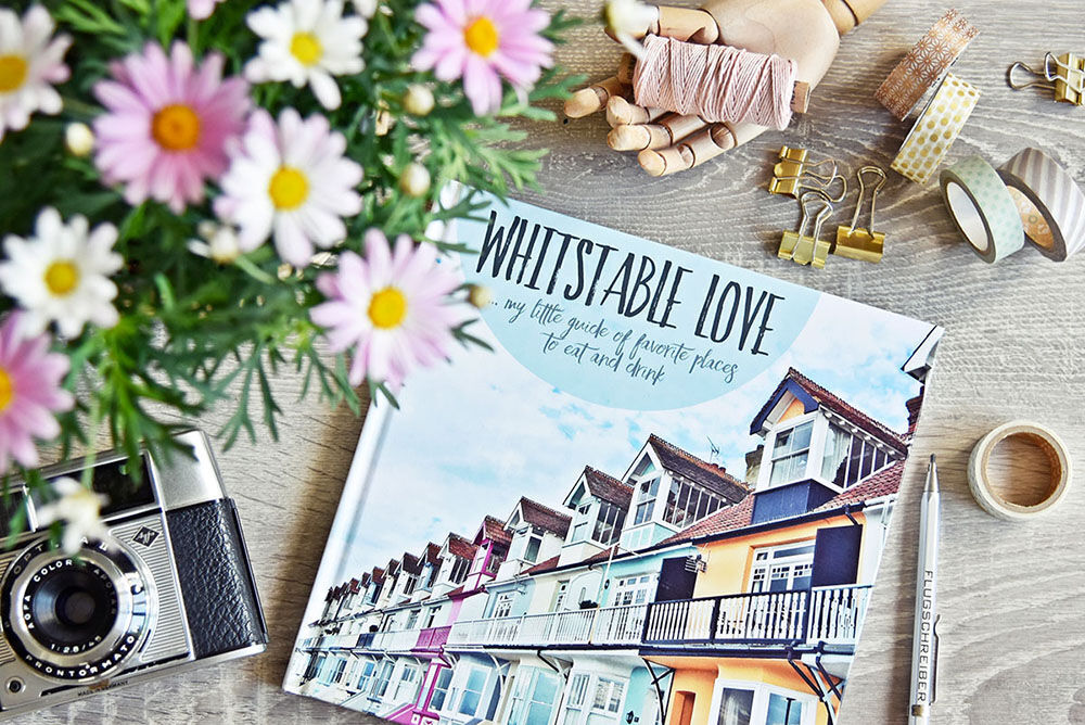 A lovingly designed photo book with colorful houses on the cover lies on a table with flowers and craft supplies.