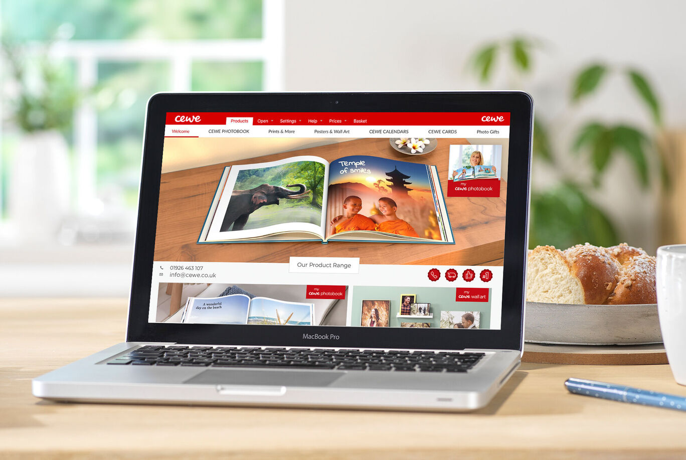 A laptop shows the CEWE FOTOBUCH website with an open photo book and images of animals and landscapes.