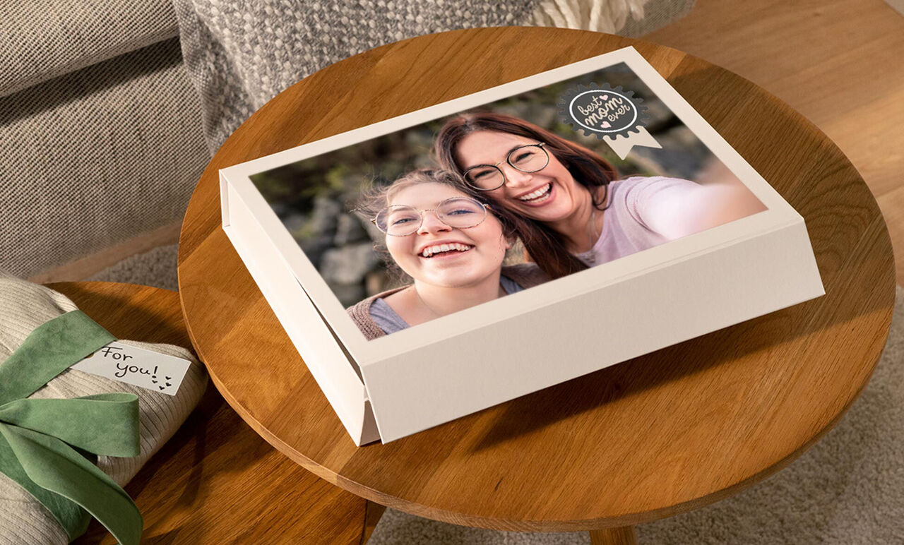 A CEWE photo book resting on a table. It shows moments of two people on the cover. A gift with a green ribbon is beside the book.