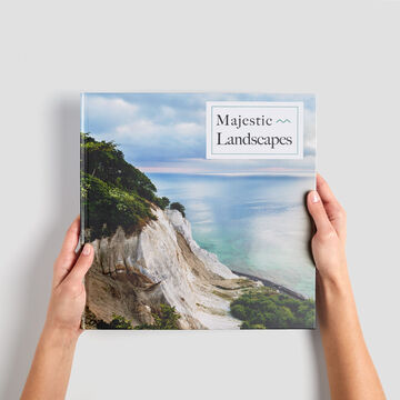 A person holds a CEWE PHOTO BOOK titled 'Majestic Landscapes' in front of a picturesque coastal landscape.