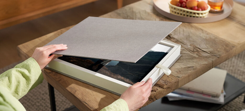 A person opens a CEWE photo book on a wooden table. Below is a picture of a natural landscape. Fruits and drinks are in the background.