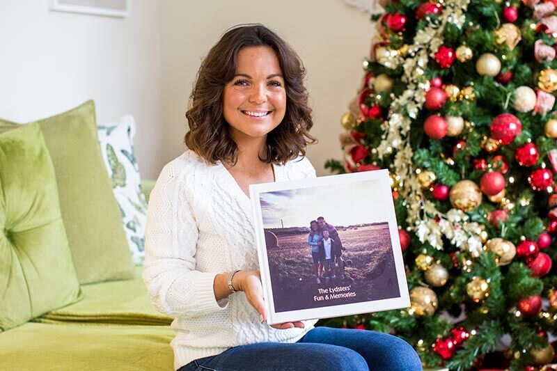 A person sits in the living room holding a photo book with family memories in front of a decorated Christmas tree.