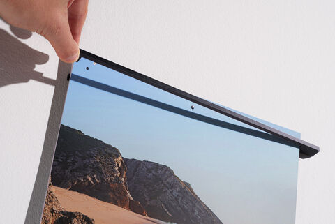 A person carefully pulls at the top edge of a wall picture to hang it. In the background, a beach with rocks and clear sky.
