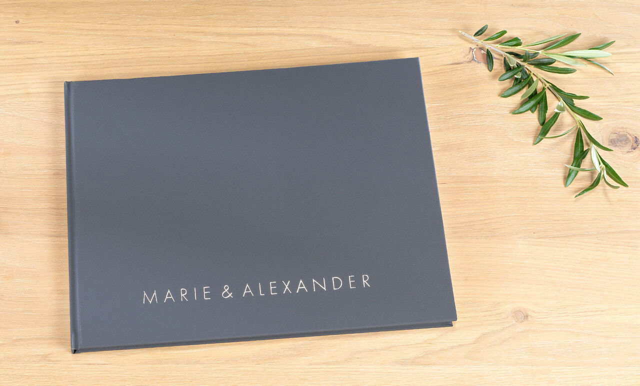 Dark photo book titled 'Marie & Alexander' on a wooden surface with an olive branch.
