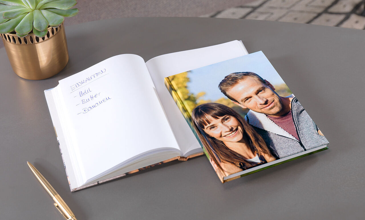 An open photo book and a calendar on a table, surrounded by a green plant arrangement