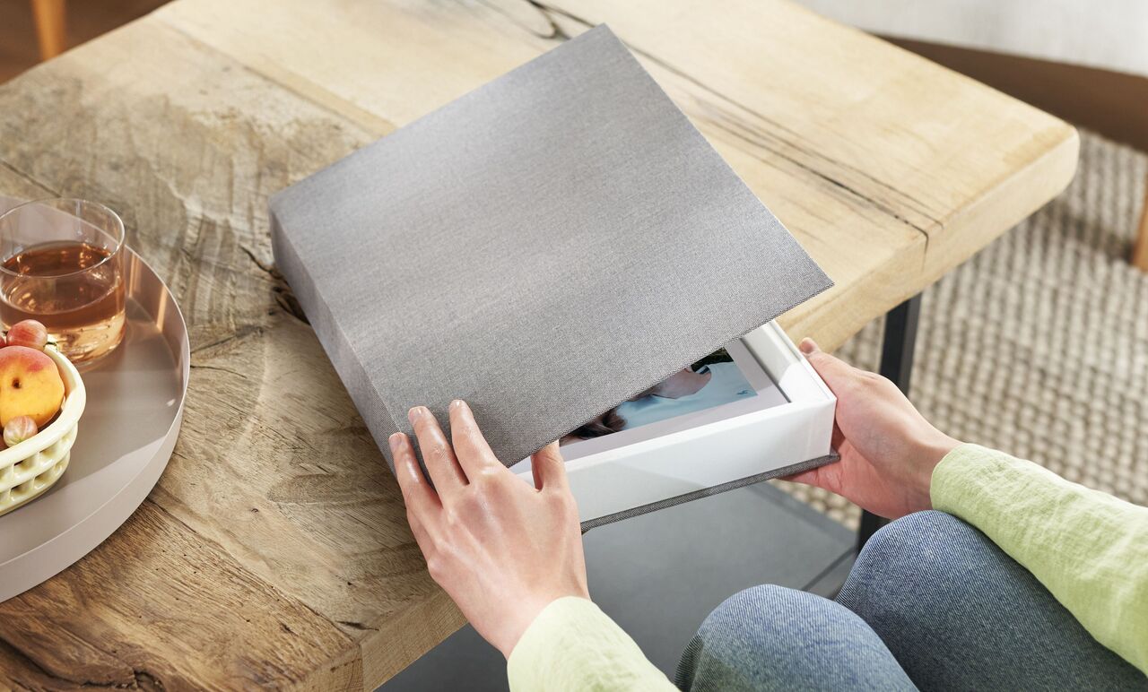 A person opens a gray CEWE photo book on a wooden table. The book is in a white box.