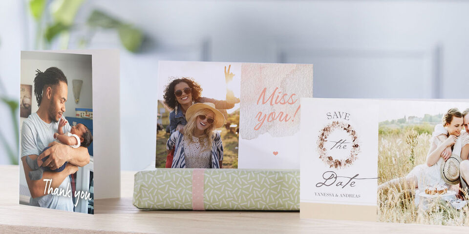 Three creative greeting cards on a table, one tall card in white with heart and text 'Miss You', next to a wedding invitation with floral wreath and 'Save the Date'