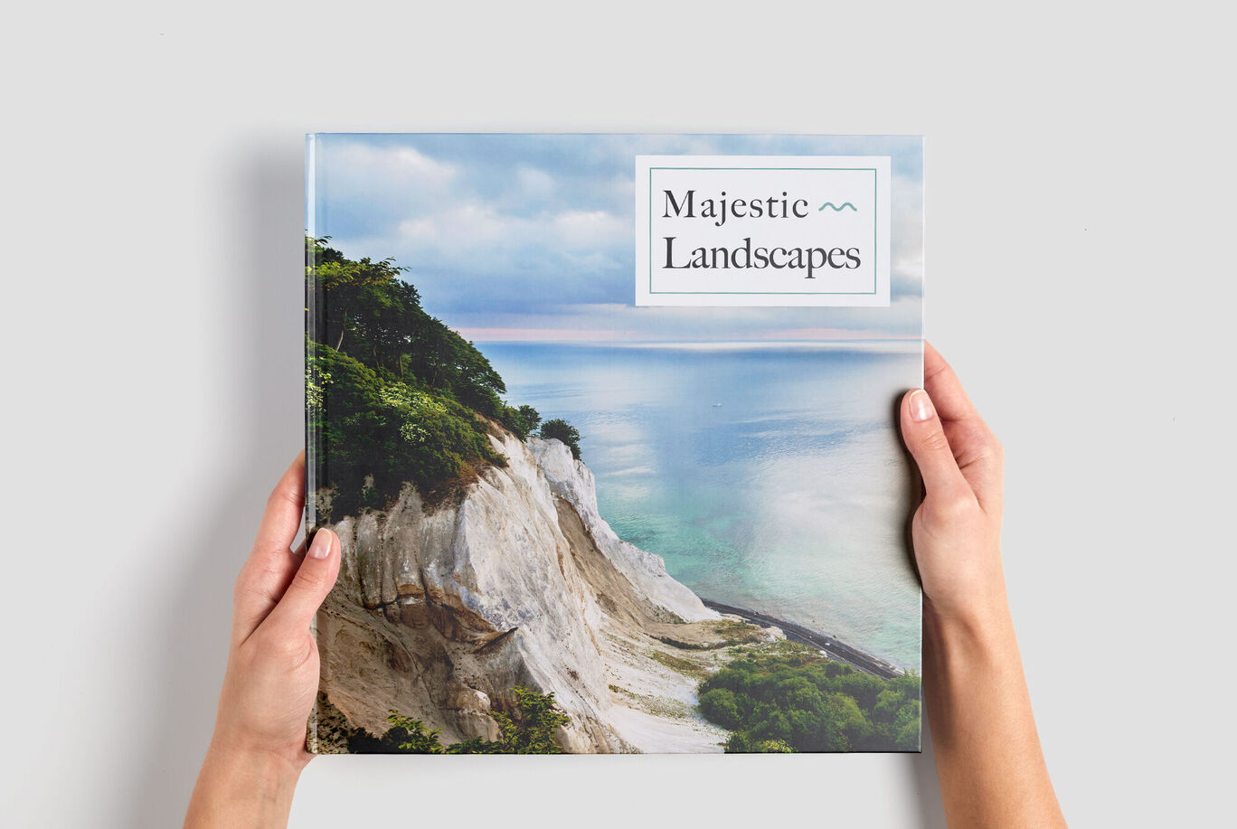 A person holds a CEWE photo book titled 'Majestic Landscapes' in front of a beautiful coastal image.