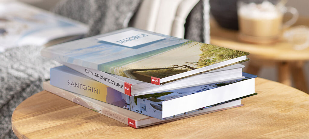 Stacked CEWE photo books with memorable moments, one shows a beach and an architectural view.
