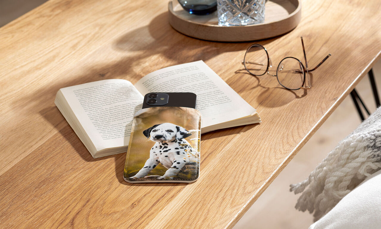 A smartphone with a dog motif lies on a table next to an open book and glasses