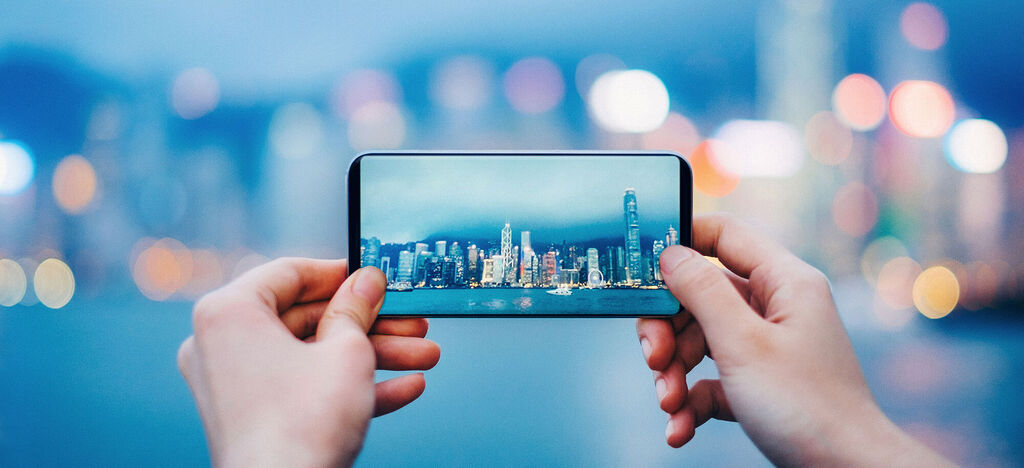 A person holds a smartphone with a picture of Hong Kong's skyline. The city lights shine beautifully at night.