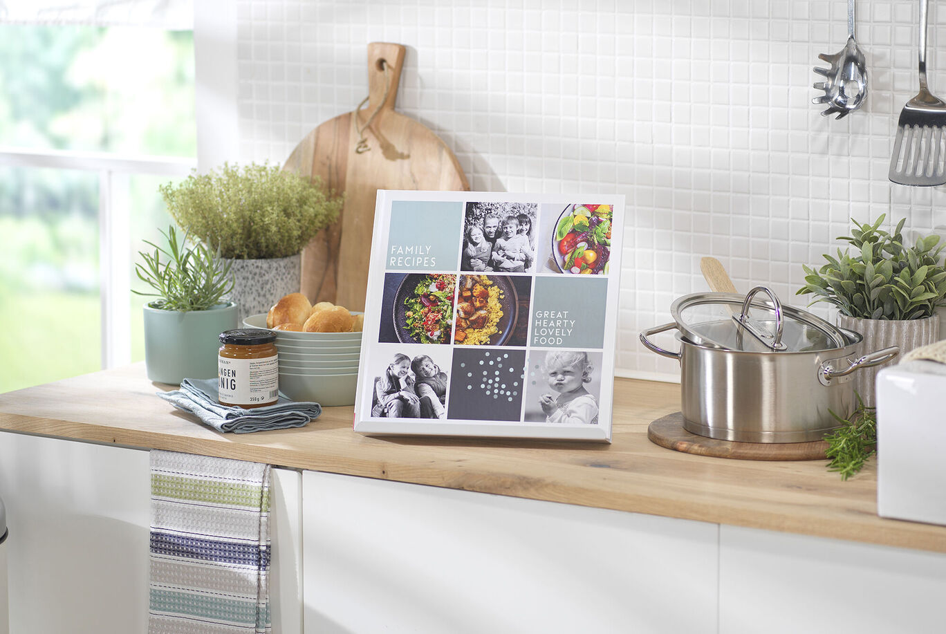 A CEWE PHOTO BOOK is on a kitchen table, surrounded by fresh herbs, bread, and a pot. Many delicious recipes are featured on the pages.