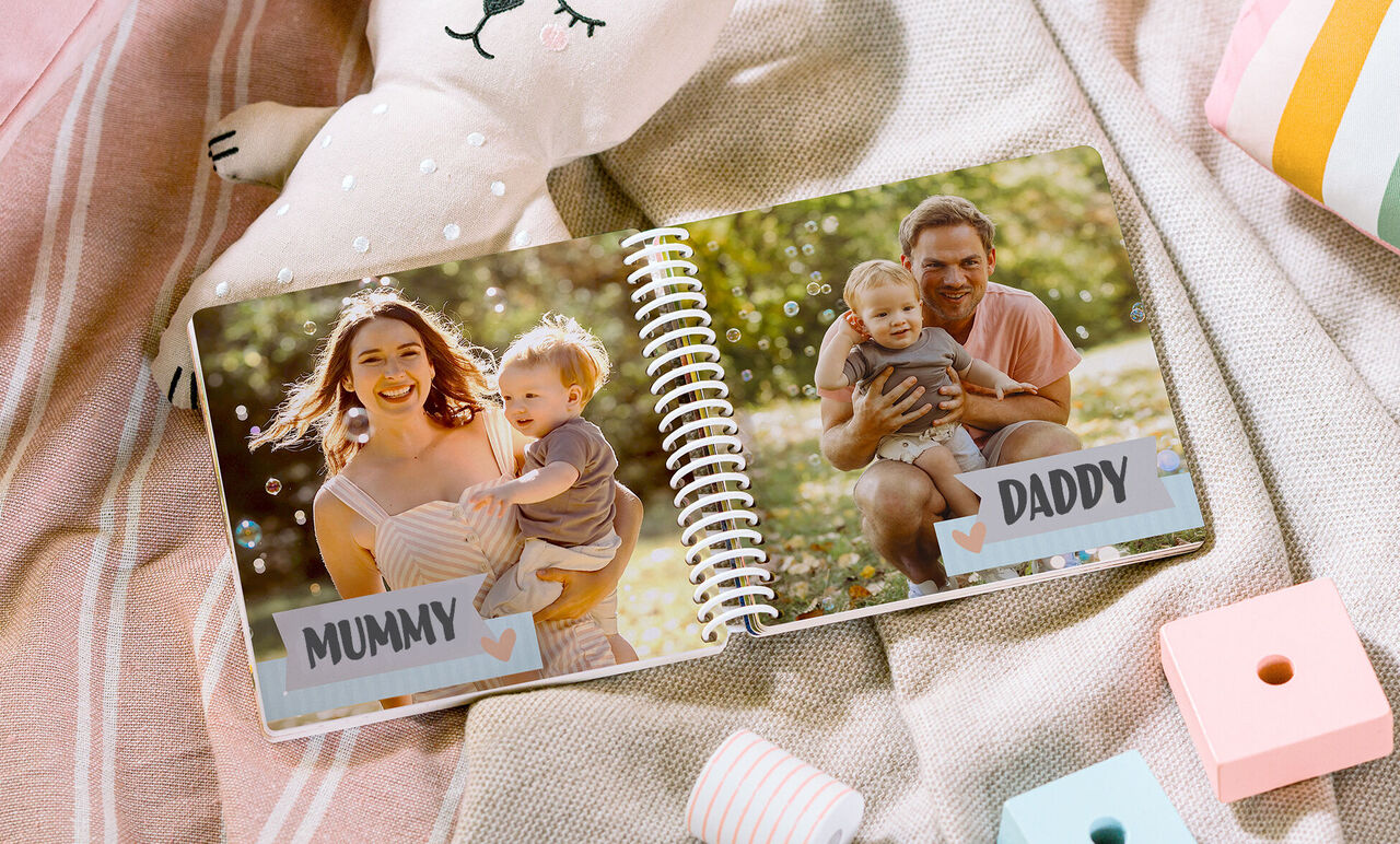 An open photobook on a cozy surface with toys. On the left, a mother with her child. On the right, a father holds his child.