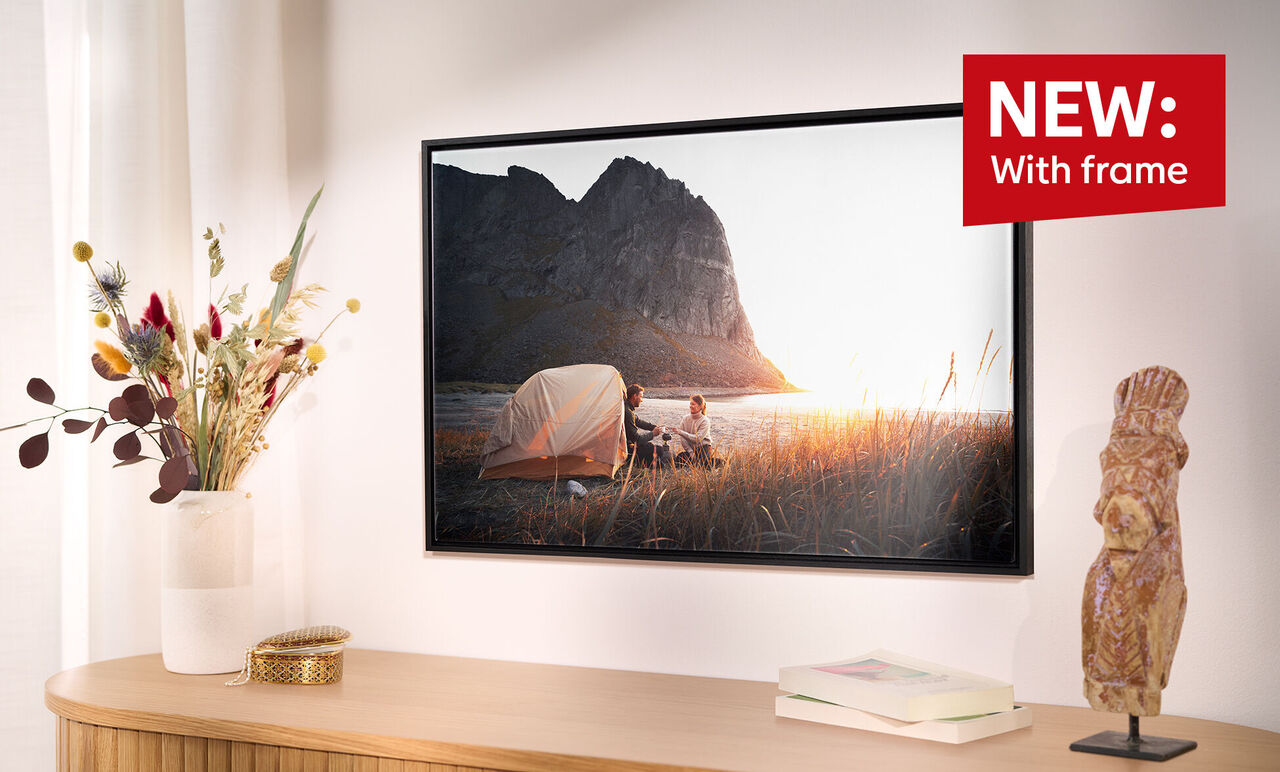 A new wall picture in an elegant frame shows a couple enjoying a romantic moment at sunset on the beach. A vase of dried flowers and a wooden figure are beside the picture.