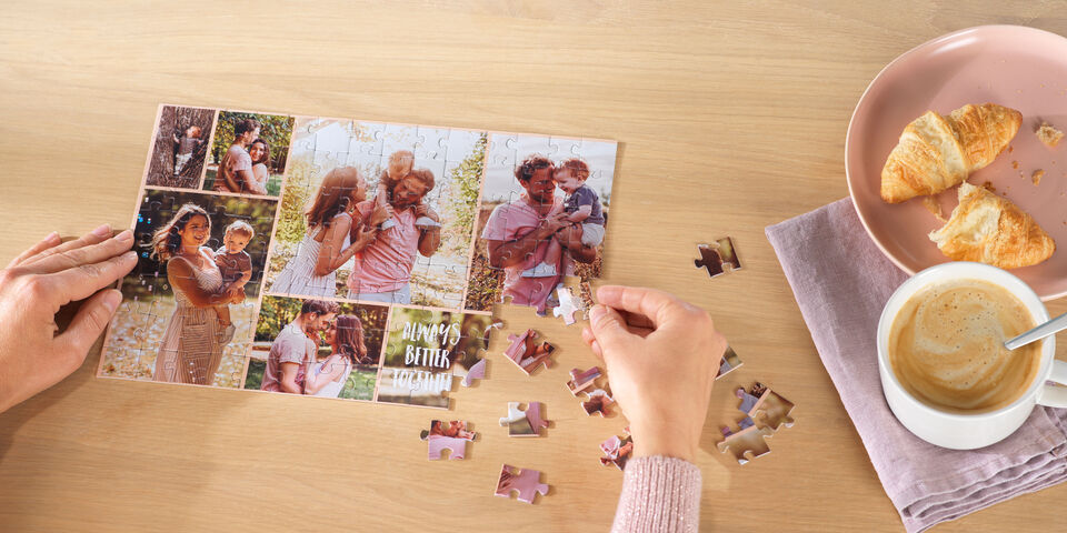 A person places a puzzle piece on a table. On the table are photos, a croissant, and a cup of coffee.
