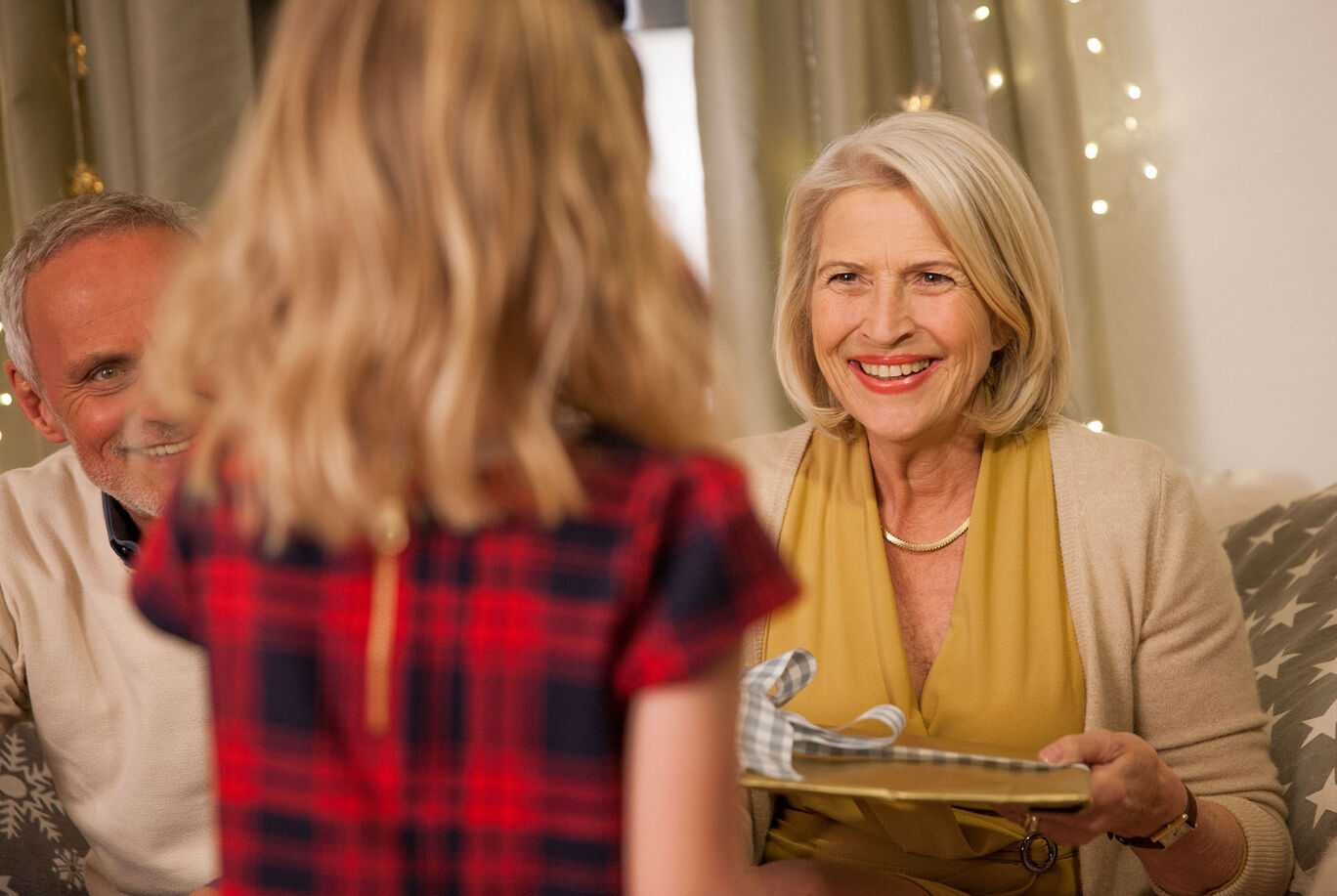 A girl hands a lovingly wrapped gift to an older woman, with a festive atmosphere in the background.