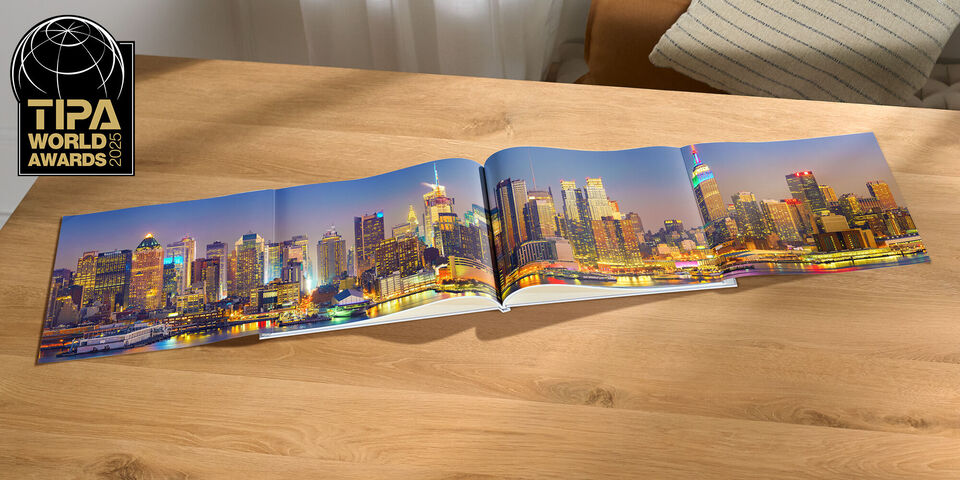 An open CEWE PHOTO BOOK on a wooden table shows a beautiful city view at night with colorful lights.