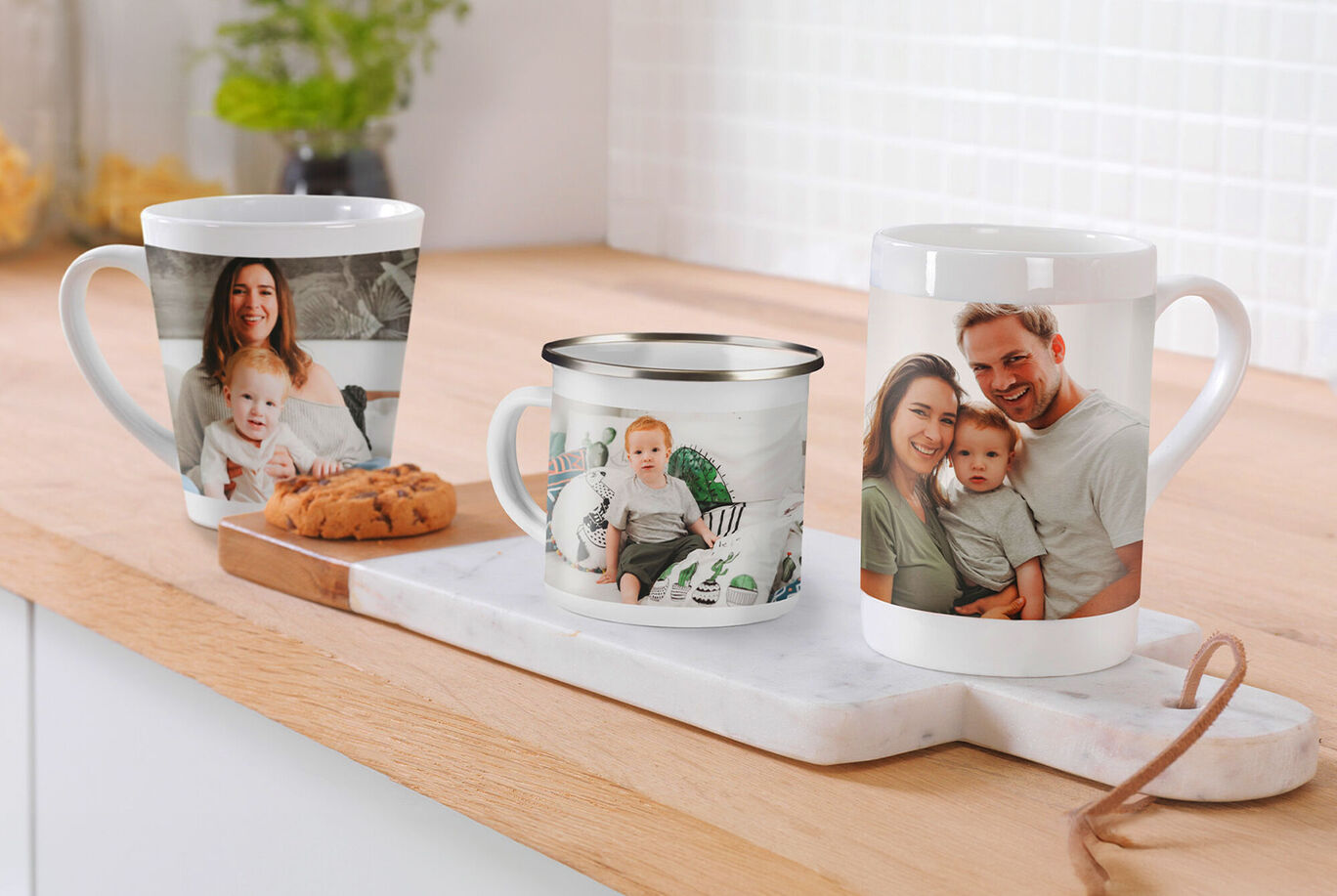 Three personalized mugs, two ceramic and one metal, placed on a wooden board. One has a photo of a small child, another shows a man and a woman. There's also a cookie on the board.