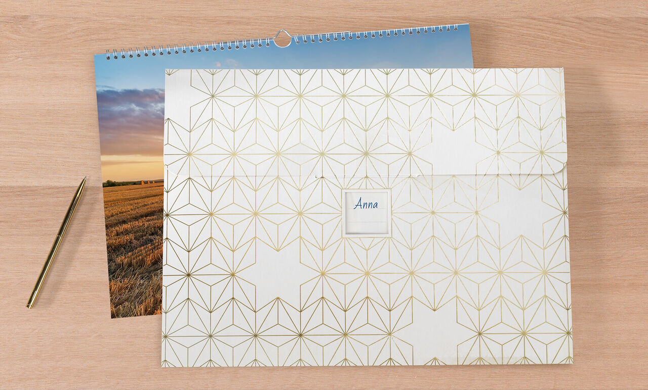 A closed CEWE photo calendar on a wooden surface, next to a photo and a gold pen.