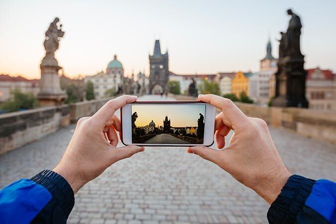 A person photographs the Charles Bridge in Prague at sunset with a smartphone. The bridge is surrounded by historic buildings.