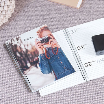 An open notebook with a photo motif on the cover showing a person with a camera. On the right, calendar sheets for planning are visible.