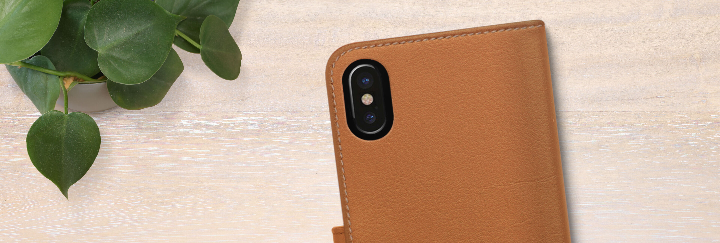 Close-up of a brown phone case on a light wooden table, accompanied by a green plant.