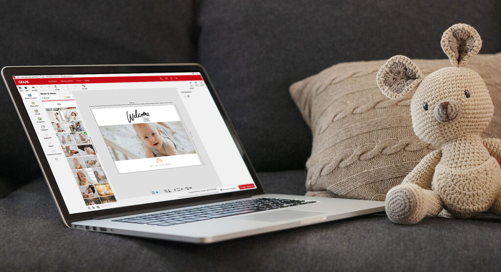 Laptop displays a design of CEWE photo book with a cute baby photo and a stuffed animal on a couch.