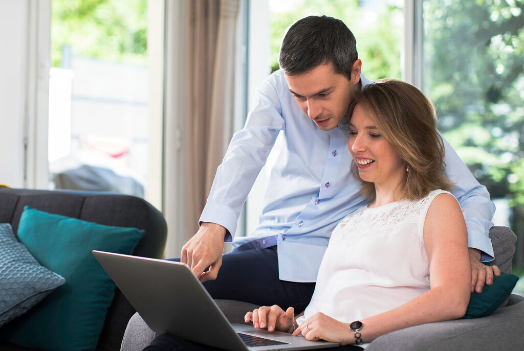 A couple sits together on a couch and looks at a laptop. They seem interested and share a friendly moment.