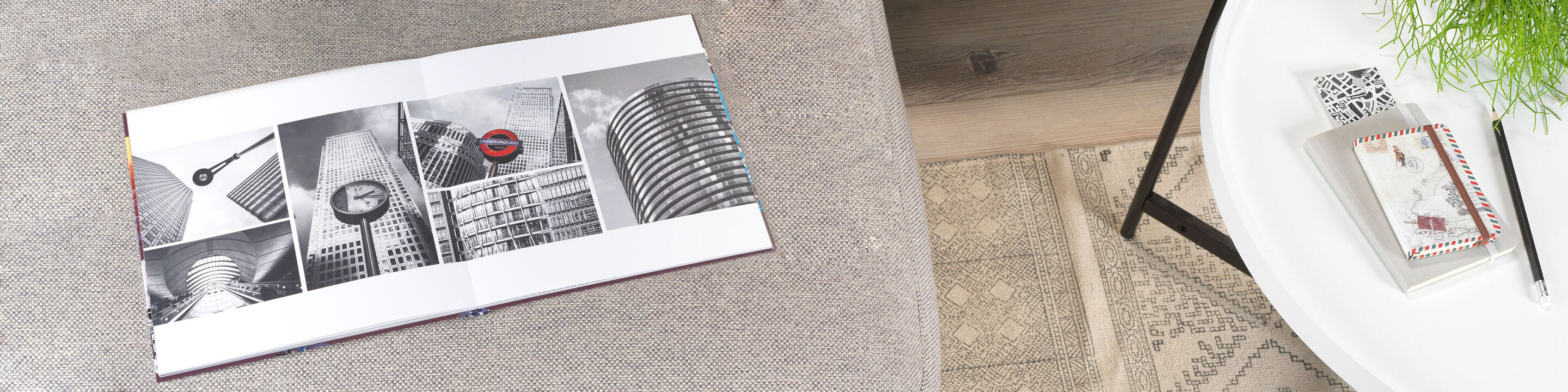 An open CEWE photo book with black and white city shots, on a table beside a notebook and a plant.