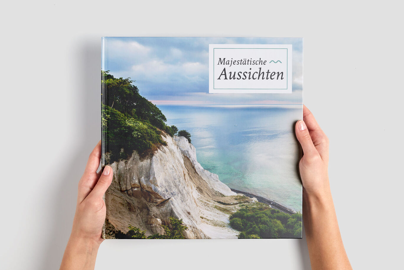 A person holds a CEWE photo book titled 'Majestic Views' in front of a beautiful coastal landscape.