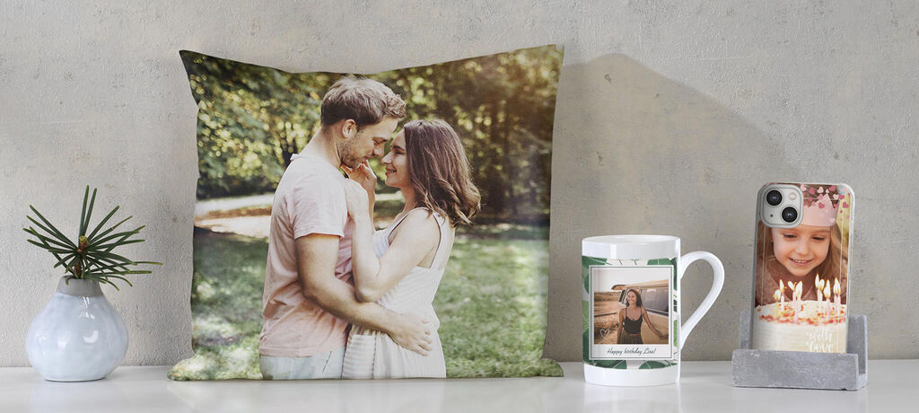 Two people embrace in front of a natural backdrop, surrounded by photo products like pillows, cups, and phone cases.