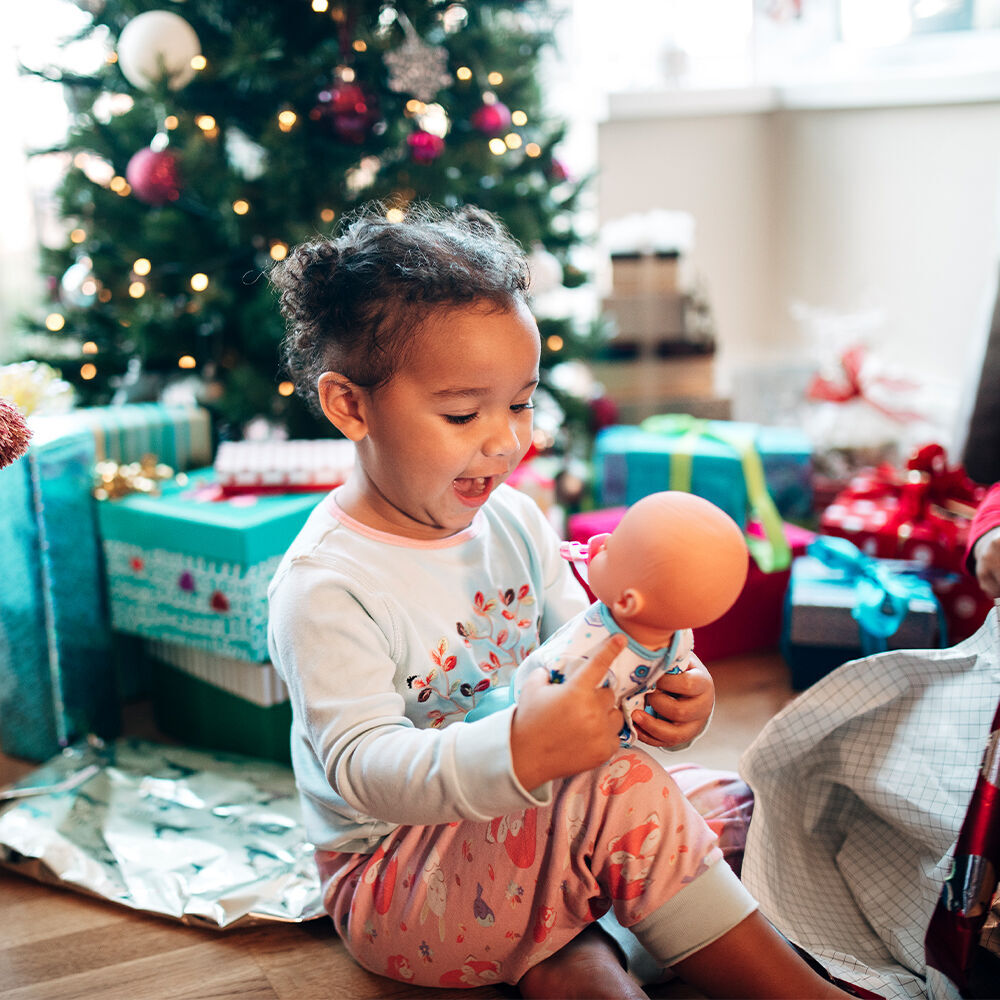 Small child sits in front of a Christmas tree and plays with a doll