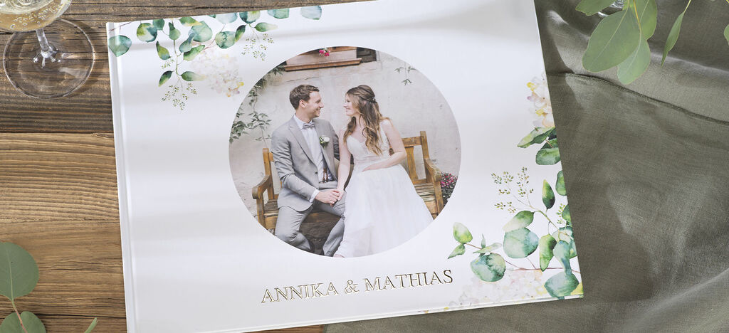A happy couple sits with a photo book on a table, surrounded by plants.