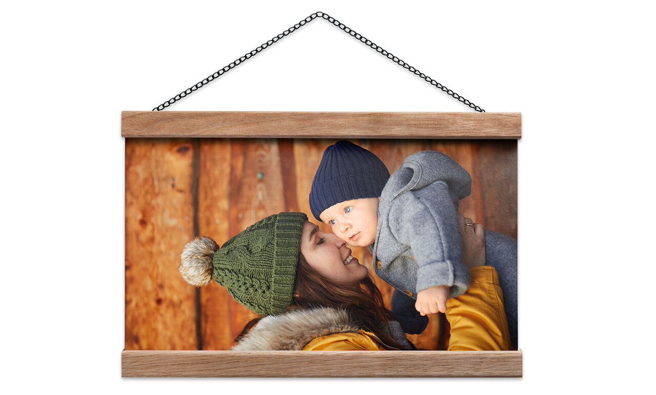 A smiling mother holds her baby in warm clothing against a wooden wall.