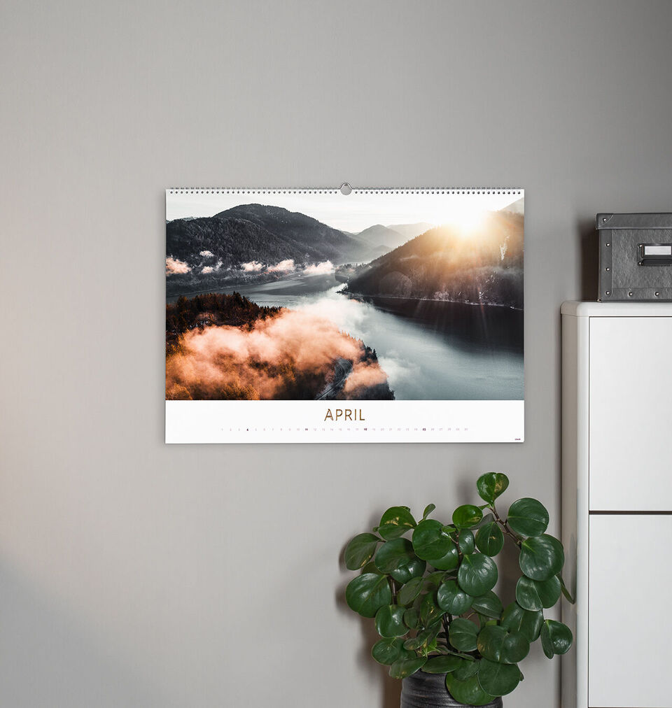 A calendar with an image of mountains and a lake in the background. The sun shines through the clouds with a plant on a black table.