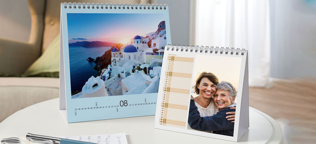 A beautiful table with two CEWE photo calendars. The left calendar shows a landscape with water and hills, while the right shows a loving photo of two people.